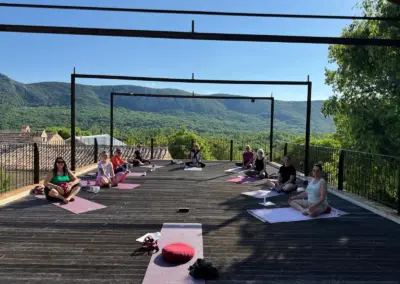Gruppe von Menschen sitzt auf Yogamatten auf einer Terrasse mit Blick auf grüne Berge.